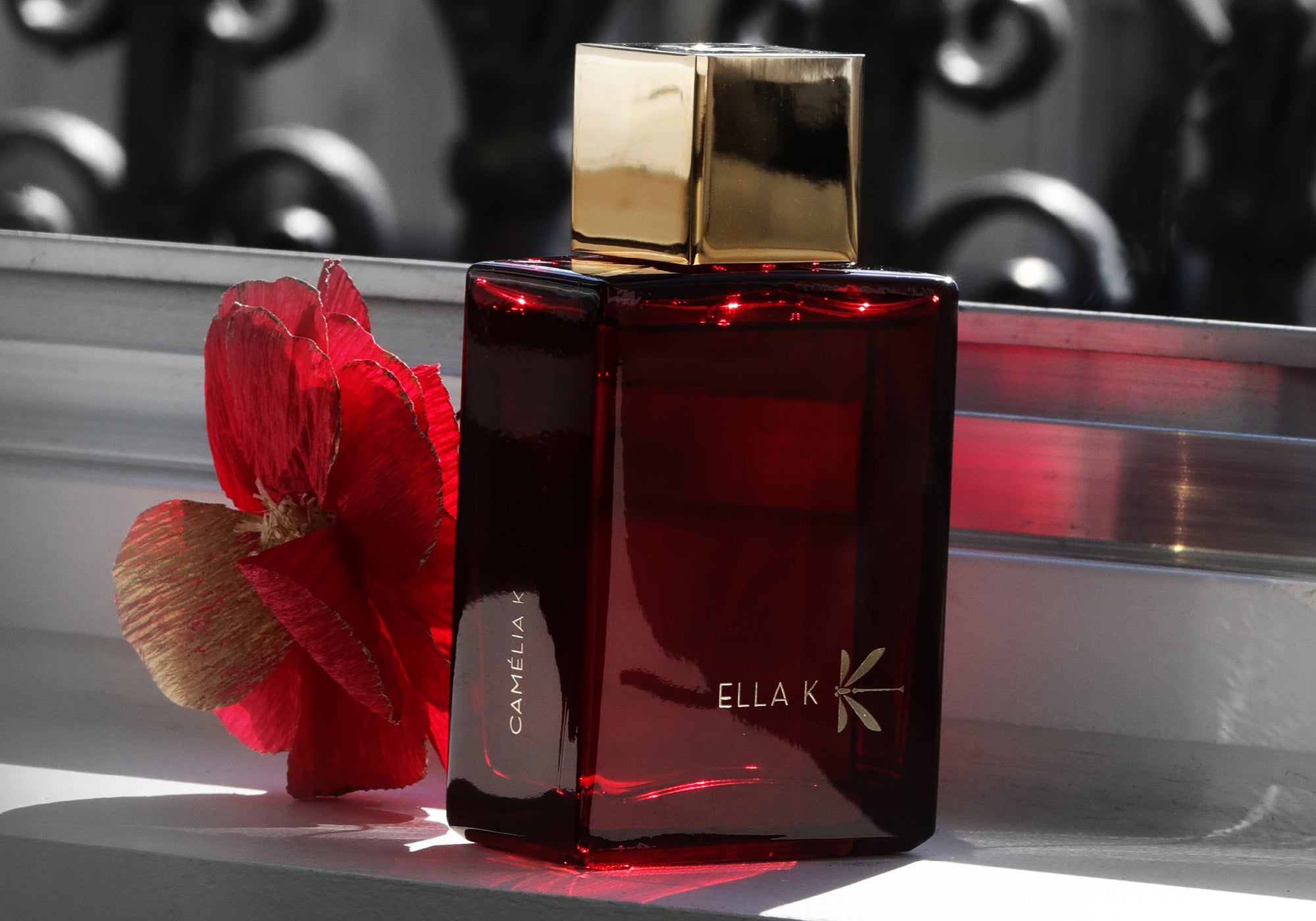 Flacon Camelia K d’Ella K avec bouchon doré sur une fenêtre, à côté d’une fleur de camélia rouge – reflets accentuant le verre rouge profond.