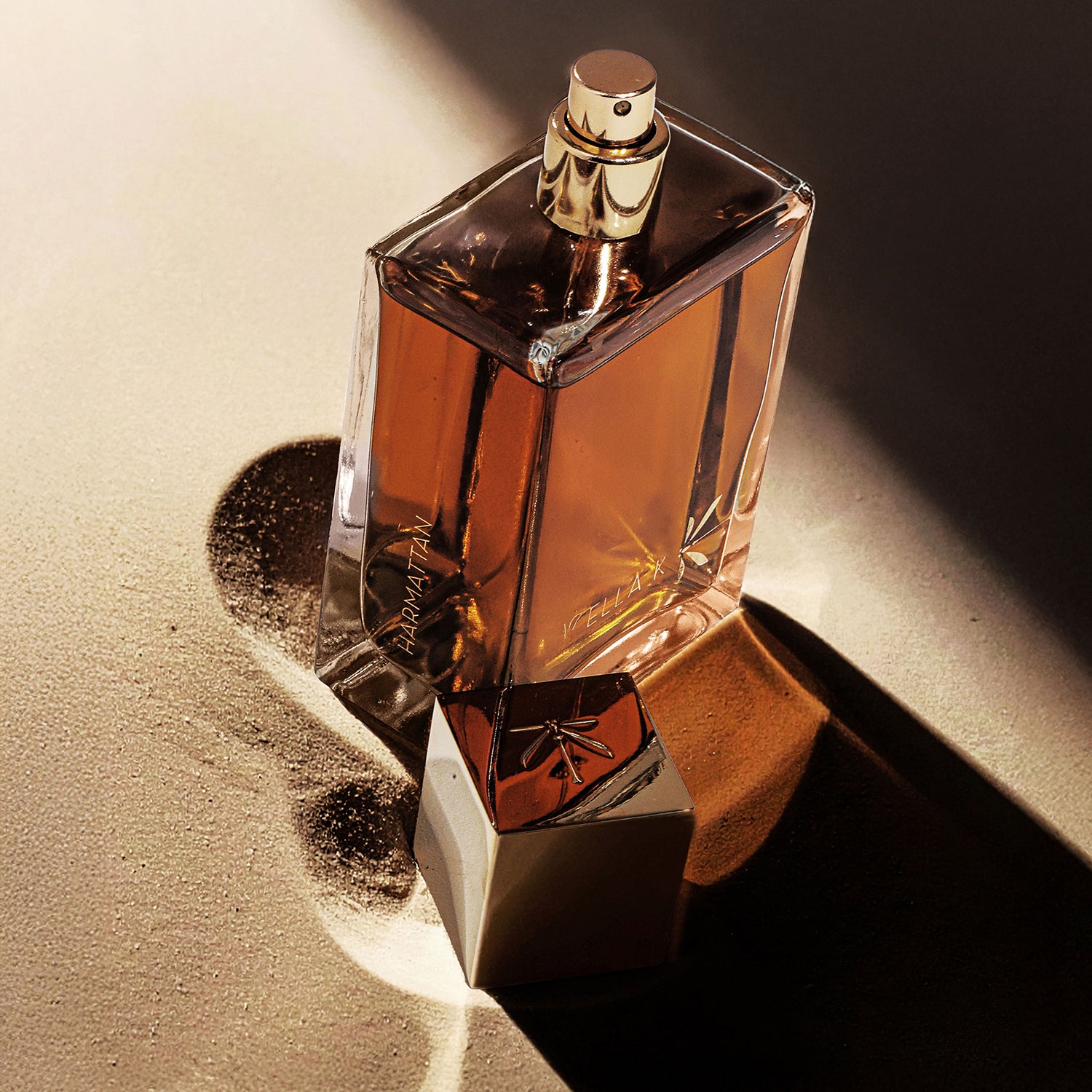 Flacon de parfum Harmattan avec capuchon doré, posé sur le sable avec jeu d’ombres – symbole de vent, désert et sensualité
