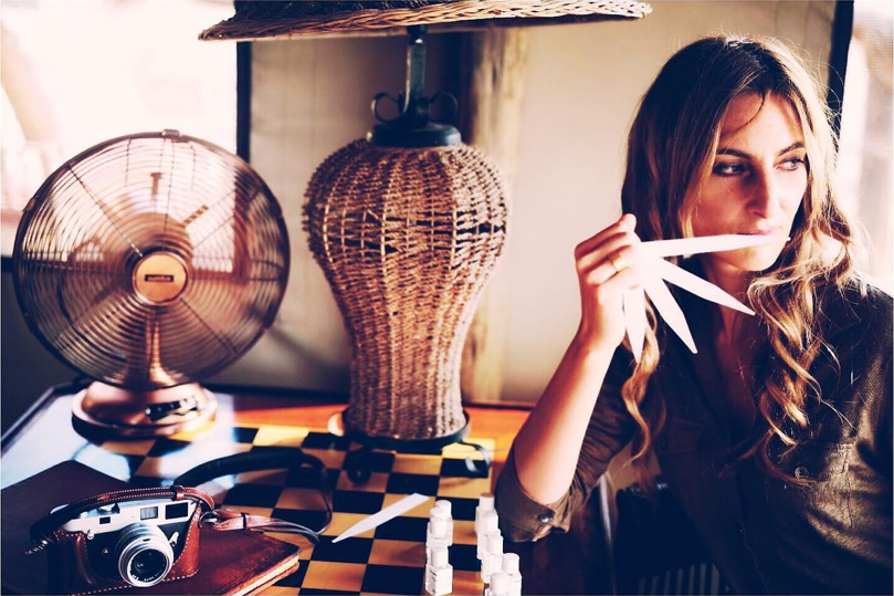 Portrait de Sonia Constant en pleine création olfactive – elle tient des touches à sentir tout en composant de nouveaux accords de parfum. À l’arrière-plan, un appareil photo vintage, un ventilateur et une lumière chaude illustrent l’univers créatif d’Ella K Parfums.