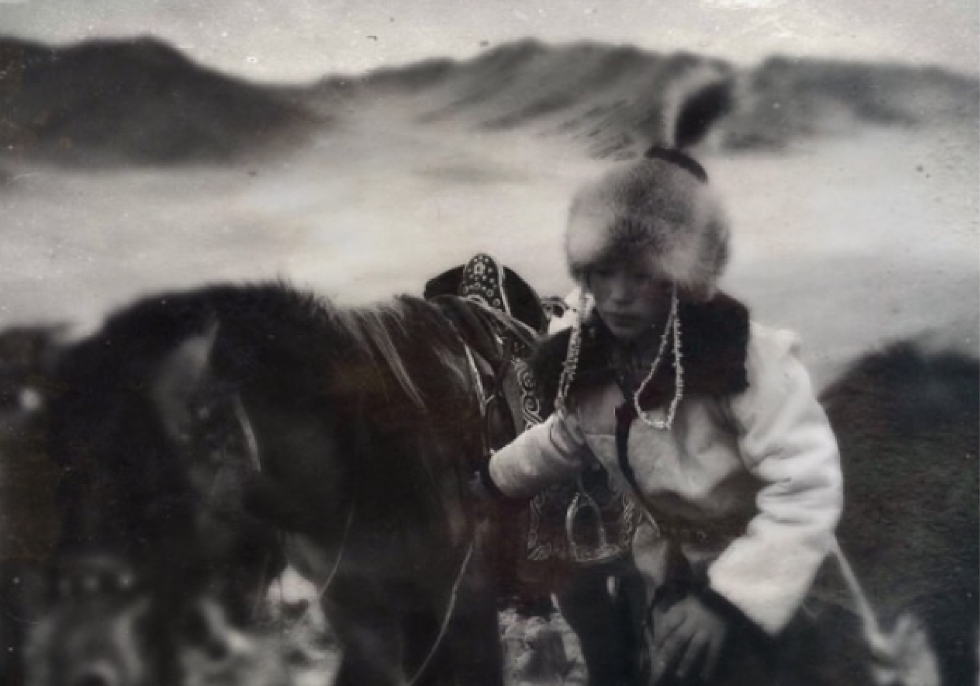 Photo noir et blanc d’un enfant nomade en tenue traditionnelle et chapeau de fourrure menant un cheval décoré dans une steppe montagneuse – symbole d’aventure et d’éloignement.