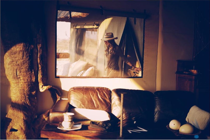 Vue à travers la tente de safari sur le reflet de Sonia Constant dans la lumière du soleil couchant – un moment de réflexion où le parfum et le souvenir se rejoignent.