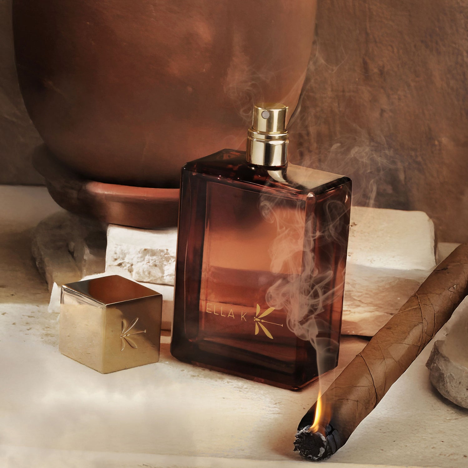 Le flacon ouvert d’Amber K posé devant un vase en terre cuite, une fine fumée de cigare souligne l’ambiance sensuelle.