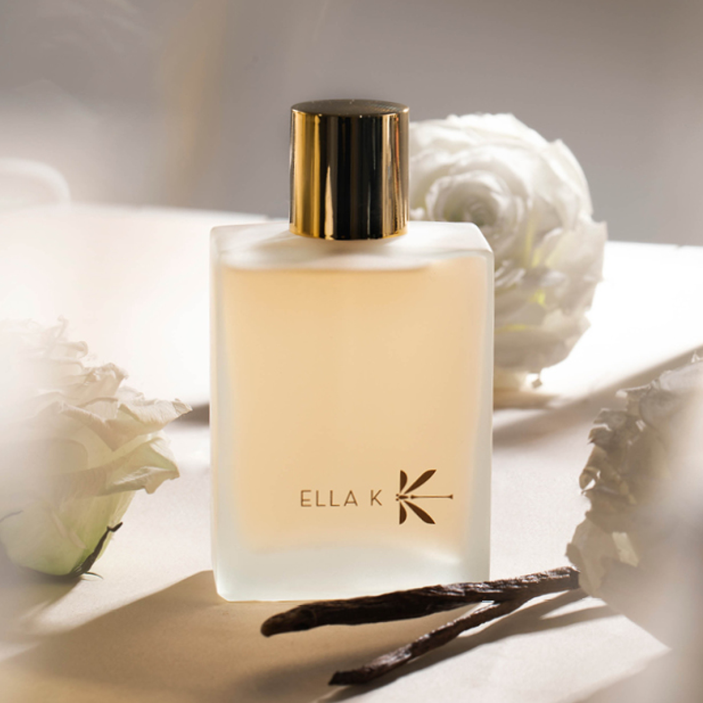 Perfume bottle of ‘Lettre de Pushkar Hairmist’ by Ella K with a gold cap, surrounded by white roses and vanilla pods, illuminated by soft light against a light background.