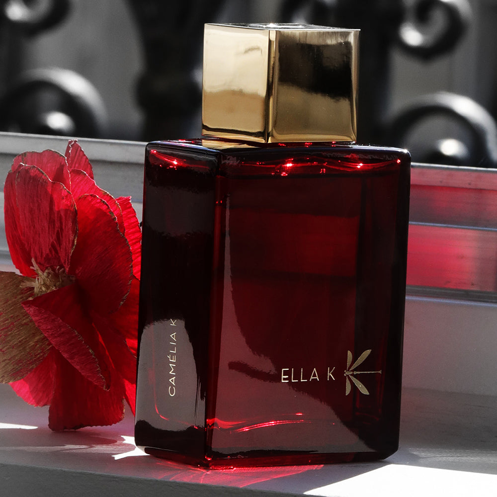 Flacon de Camélia K – rouge rubis avec bouchon doré – posé sur un rebord de fenêtre à côté d’une fleur rouge, dans une mise en scène lumineuse et élégante.