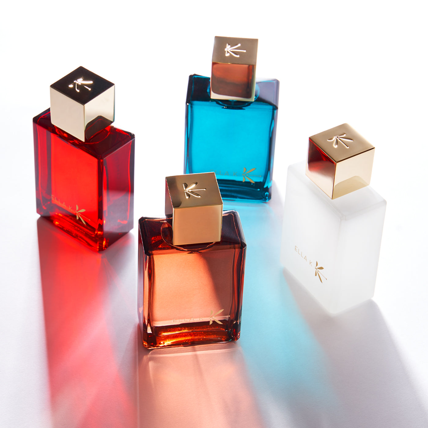 Quatre flacons de la collection K d’Ella K en couleurs vives – rouge, ambre, bleu, blanc – bouchons dorés, disposés artistiquement sur fond clair avec reflets colorés.