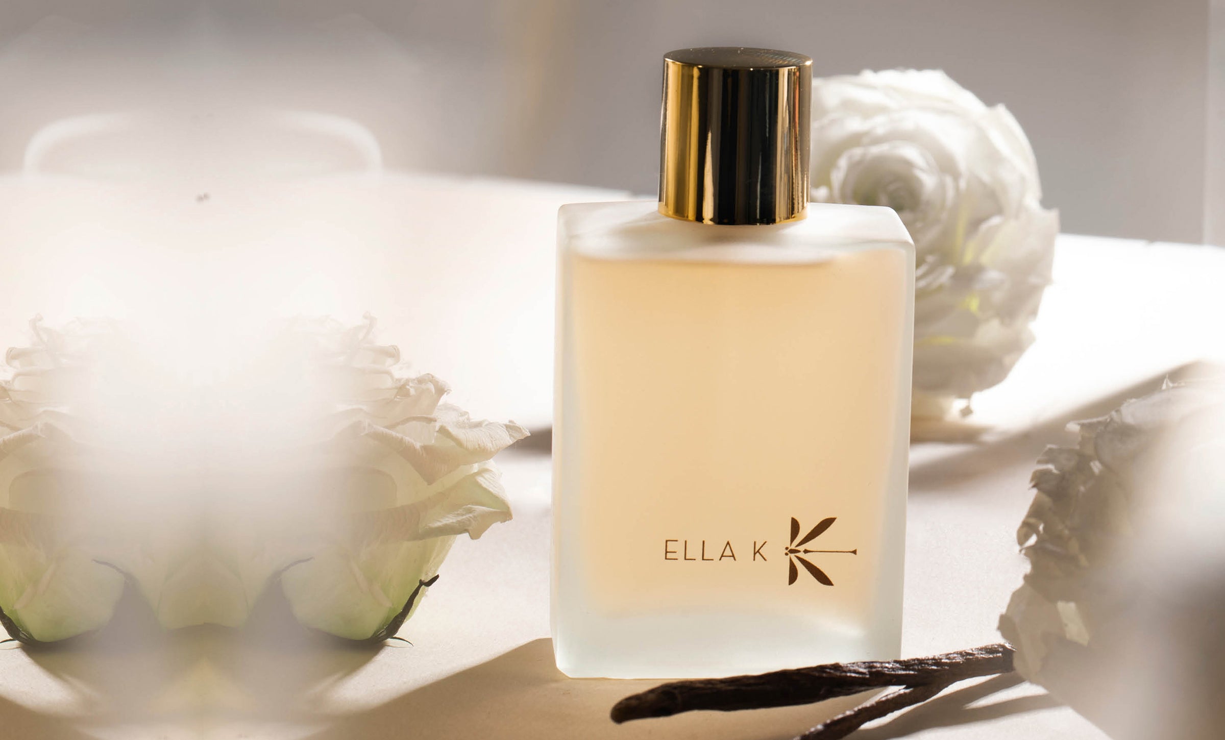 Flacon « Lettre de Pushkar Hair Mist » d’Ella K Parfums en verre givré, bouchon doré, posé sur fond clair à côté de fleurs blanches et d’une gousse de vanille.