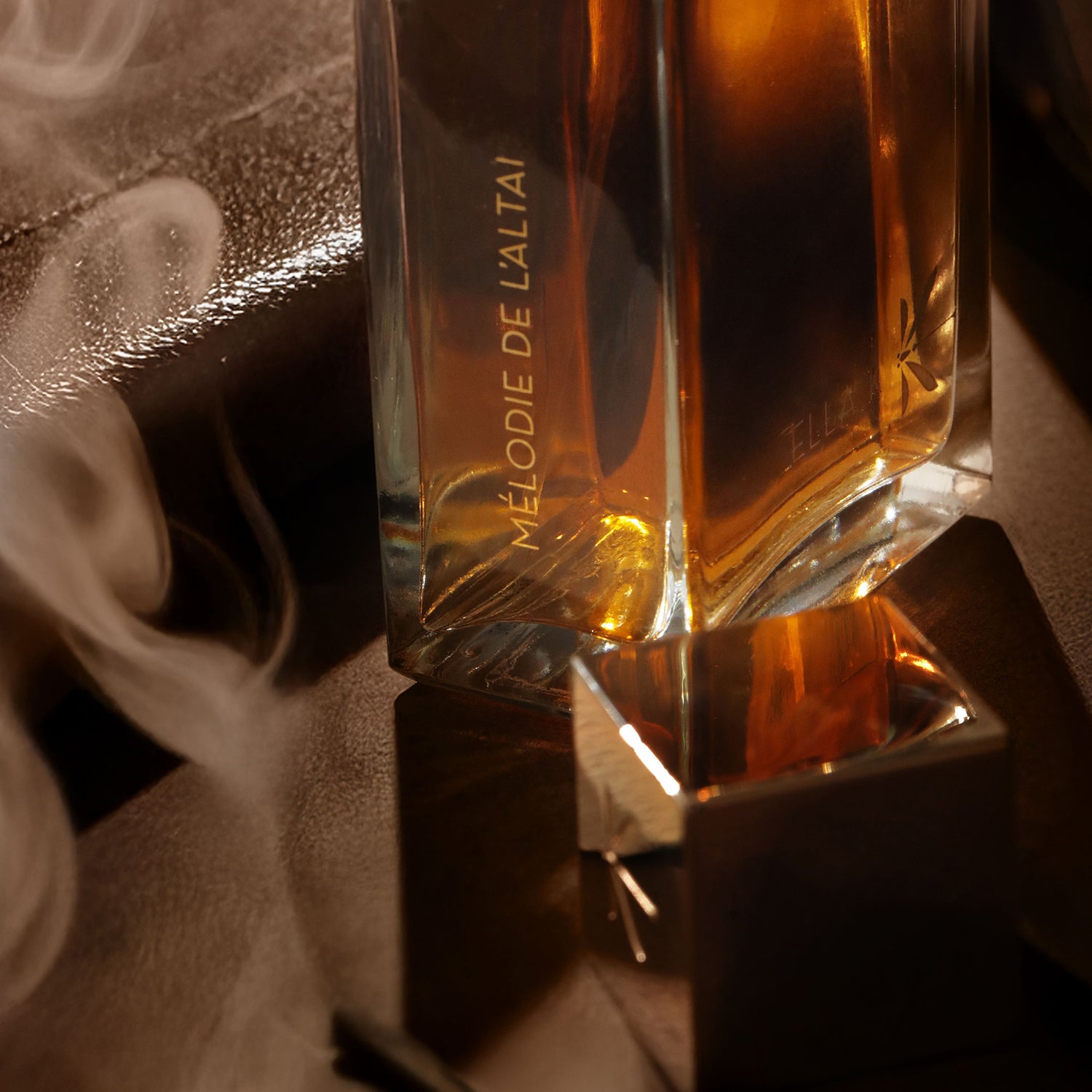Gros plan sur le flacon de « Mélodie de l’Altaï » avec le nom du parfum inscrit sur le verre. Le bouchon doré repose au premier plan sur une surface sombre, enveloppé d’un léger nuage de vapeur ou de fumée.