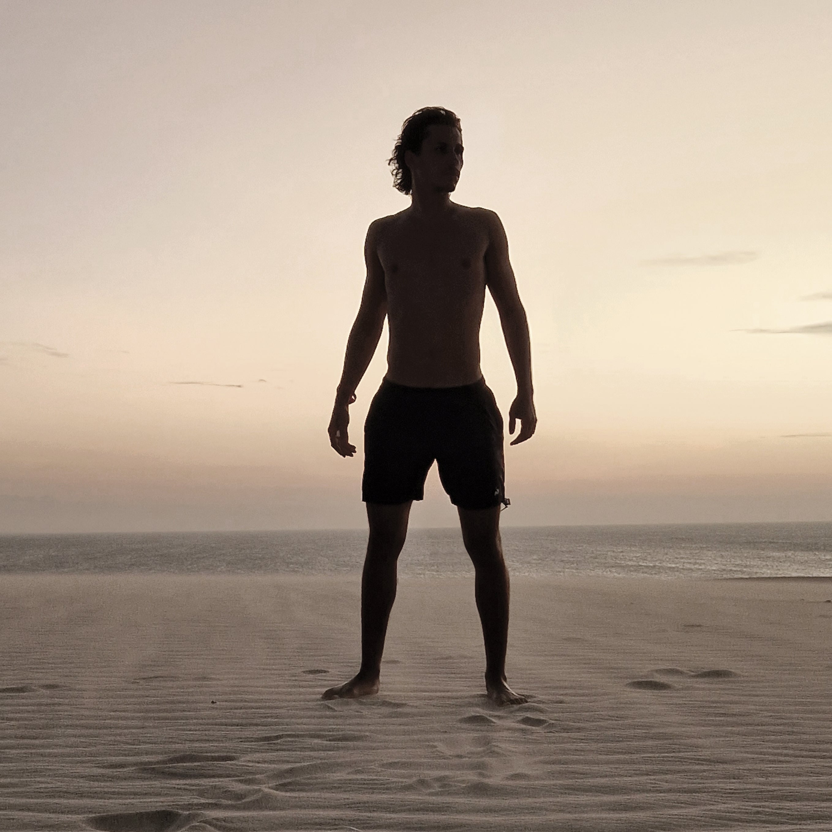 Un homme athlétique se tient seul sur une dune au coucher du soleil, face à l’océan – une image de force naturelle et de sensualité libre, reflet du parfum Musc K.