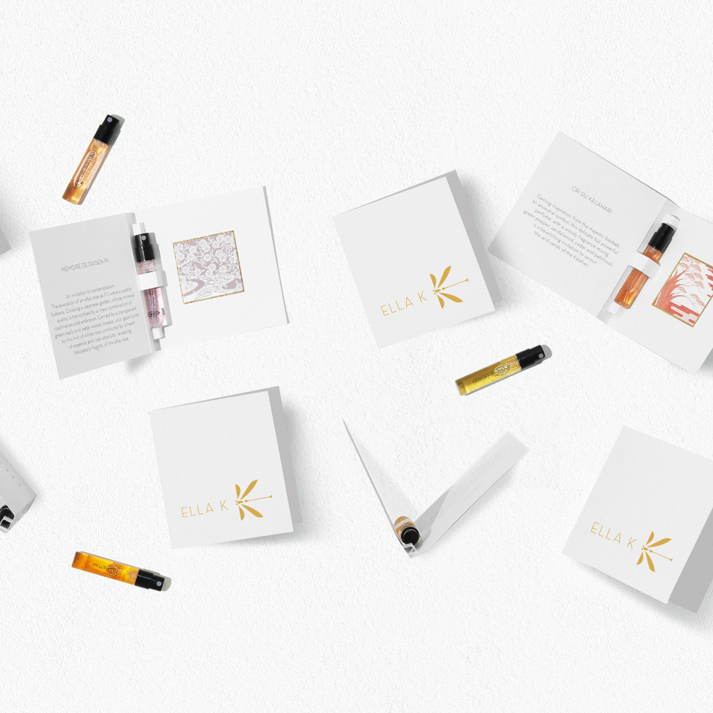 Diverses échantillons de parfum d’Ella K en petits flacons, accompagnés d’élégants livrets blancs au logo doré, disposés à plat sur fond blanc.