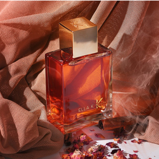 Le flacon du parfum « Lettre de Pushkar » d’Ella K devant un tissu cuivré, entouré de pétales de rose, d’un bâton de cannelle et d’un voile de fumée – une fragrance inspirée de la spiritualité sensuelle du Rajasthan.